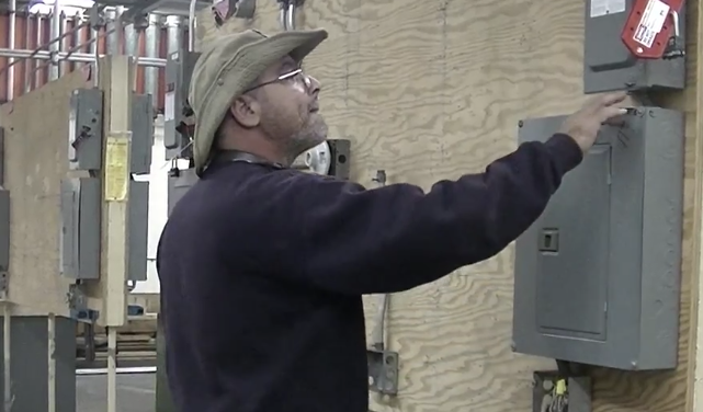 Man in cap and glasses reaching toward an electrical panel on a brick wall