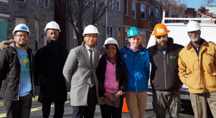 Eight people wearing hard hats stand together on a city street during a solar installation project benefiting homeowners