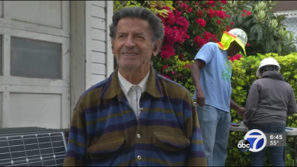 Man in striped cardigan smiles while solar installers work on roof behind him, ABC7 news broadcast
