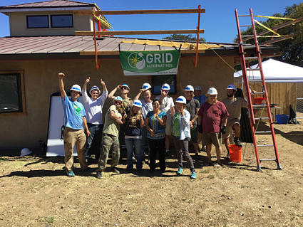Group of people in hard hats standing in front of a partially constructed home with GRID banner and solar installation scaffolding