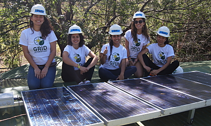 Five women in hard hats and GRID shirts examine solar panels outdoors
