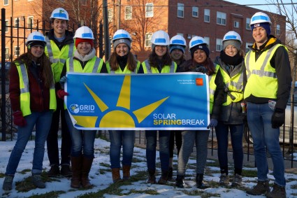 Group of volunteers in hard hats and safety vests holding Solar Spring Break banner outdoors in snow