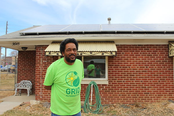 Man in green GRID shirt holds garden hose in front of brick house with solar panels on roof