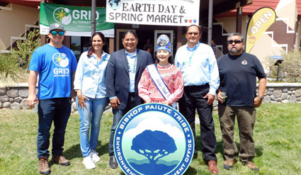 Six people stand with Earth Day & Spring Market signs and a Bishop Paiute Tribe seal banner celebrating solar system completion