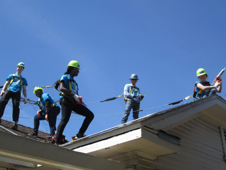 Group of youth in safety gear practicing rope work on a roofline during a solar installation training activity