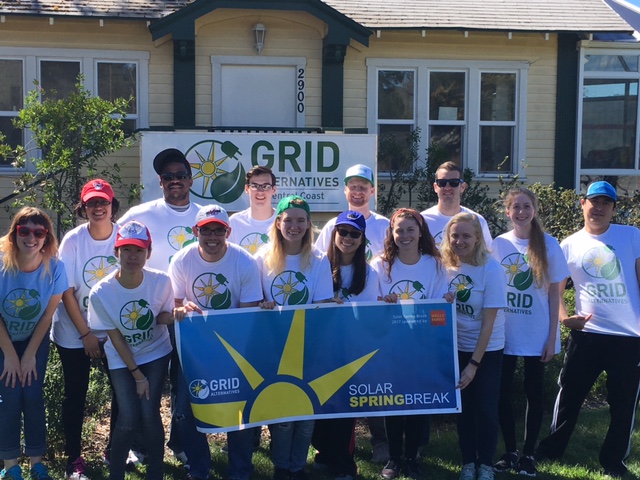 Group of volunteers in GRID Alternatives shirts holding a Solar Spring Break banner in front of a house