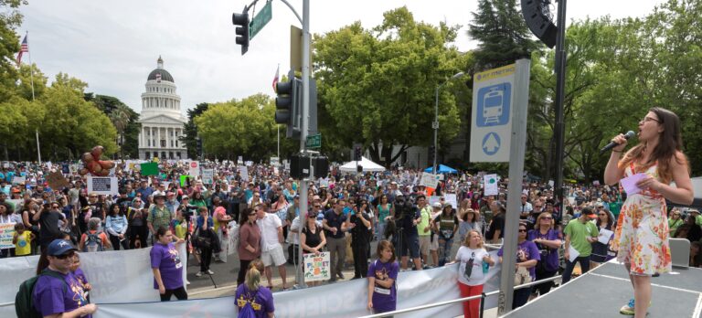 Large outdoor rally with hundreds of people holding signs in front of California State Capitol building