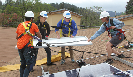 Five workers in hard hats install solar panels on a residential roof during a community green energy project