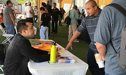 Man at table speaking with visitor at indoor career fair event with crowd in background