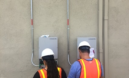 Two workers in safety gear and vests inspect wall-mounted electrical equipment during GRID workforce training