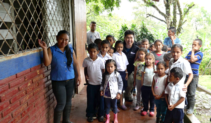 Group of children and adults gathered outdoors under a covered structure with brick walls and trees in background