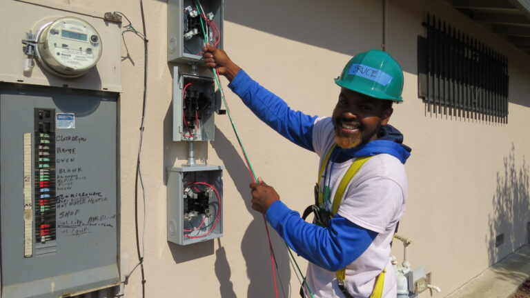 Electrician in safety gear works on residential electrical panel installation on building exterior
