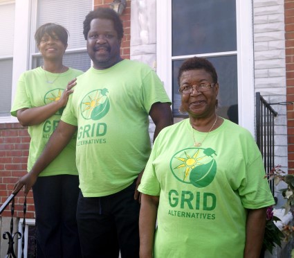 Three people wearing bright green GRID Alternatives t-shirts stand on brick house steps, promoting solar energy initiative