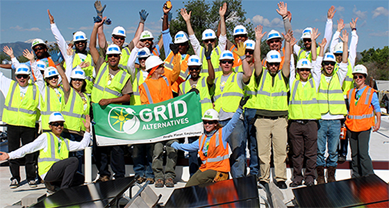 Large group of construction workers in safety gear holding GRID Alternatives banner with arms raised celebrating