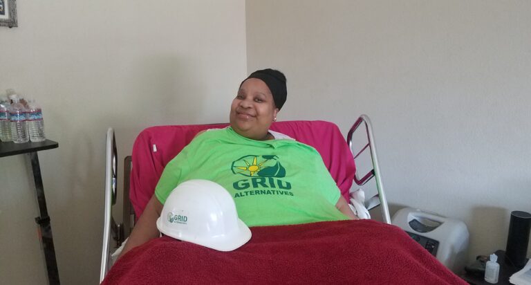 Person in green GRID Alternatives shirt holding white hard hat while reclining on red chair