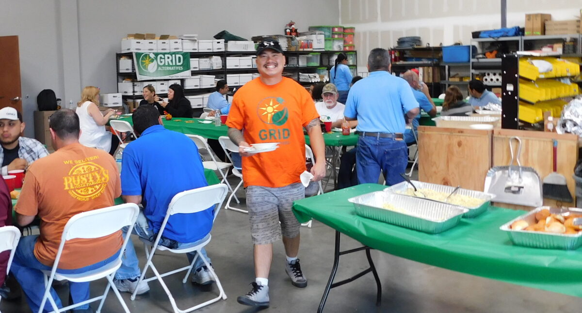 Volunteers gather in a warehouse at green tables, with a smiling volunteer in an orange GRID shirt at the center of an active food distribution event
