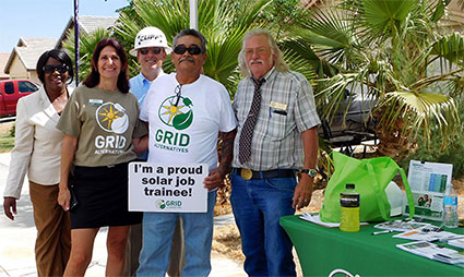 Five people at outdoor event with "I'm a proud solar trainee" sign and GRID program materials on table