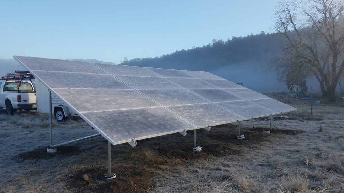 Large solar panel array mounted on ground supports with white utility vehicle nearby in rural hillside setting