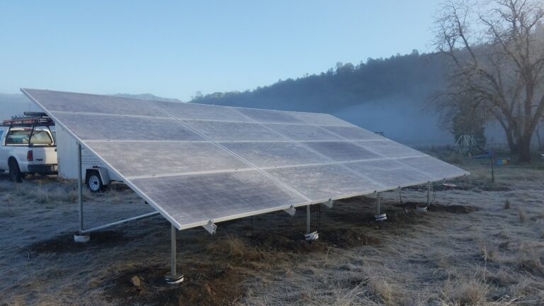 Large solar panel array mounted on ground supports with white utility vehicle nearby in rural hillside setting