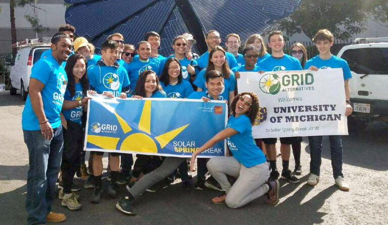 Group of volunteers in blue Grid Alternatives shirts holding banners welcoming University of Michigan to Solar Spring Break 2018