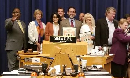 Seven people stand at a wooden podium labeled "Sheila Kuehl" during a county supervisors' awards ceremony