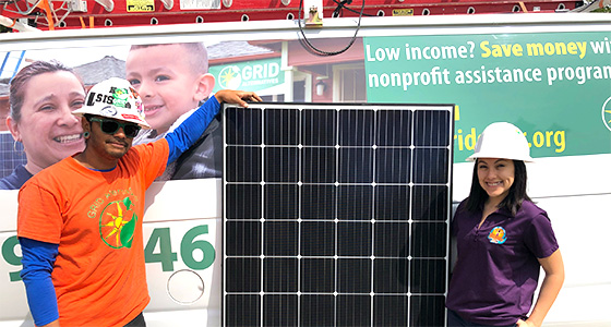 Two people posing with a large solar panel, promoting a community solar initiative with nonprofit assistance program signage visible
