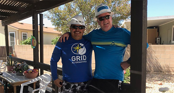 Two men wearing hats and sunglasses smile together under a patio, one wearing a "Grid Alternatives" shirt