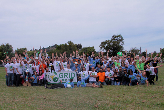 Large group of people in colorful shirts holding "GRID Alternatives" banner at outdoor event, arms raised in celebration