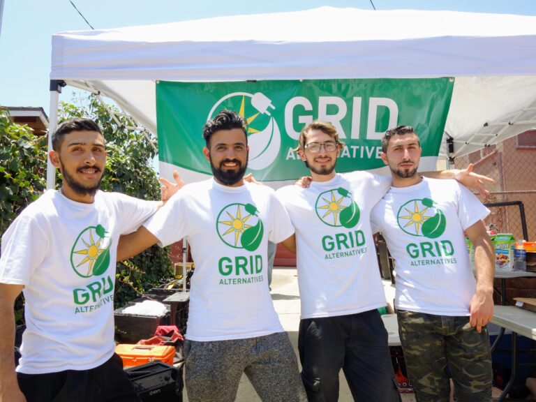 Four men wearing GRID Alternatives white t-shirts pose together under a tent at an outdoor event