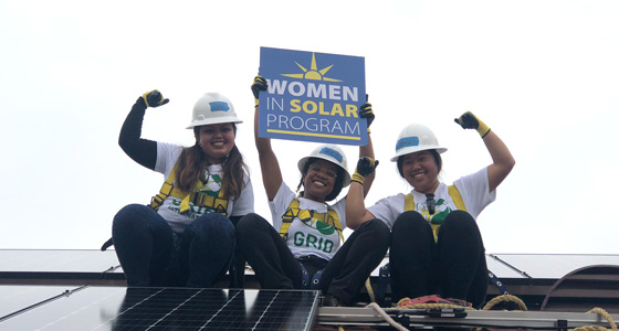 Three women in hard hats celebrating on solar panels with "Women in Solar Program" sign