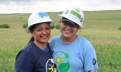 Two women in white hard hats and green solar energy logo shirts smiling in a prairie field