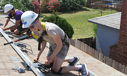 Three young people in hard hats installing solar panels on a residential rooftop during a sunny day