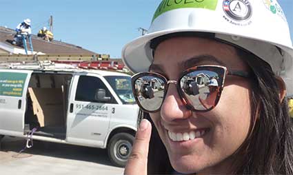 Woman in hard hat and sunglasses smiling at camera with work van and construction site behind her