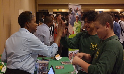 Man in blue shirt speaking with two young people at a job fair table with brochures and materials