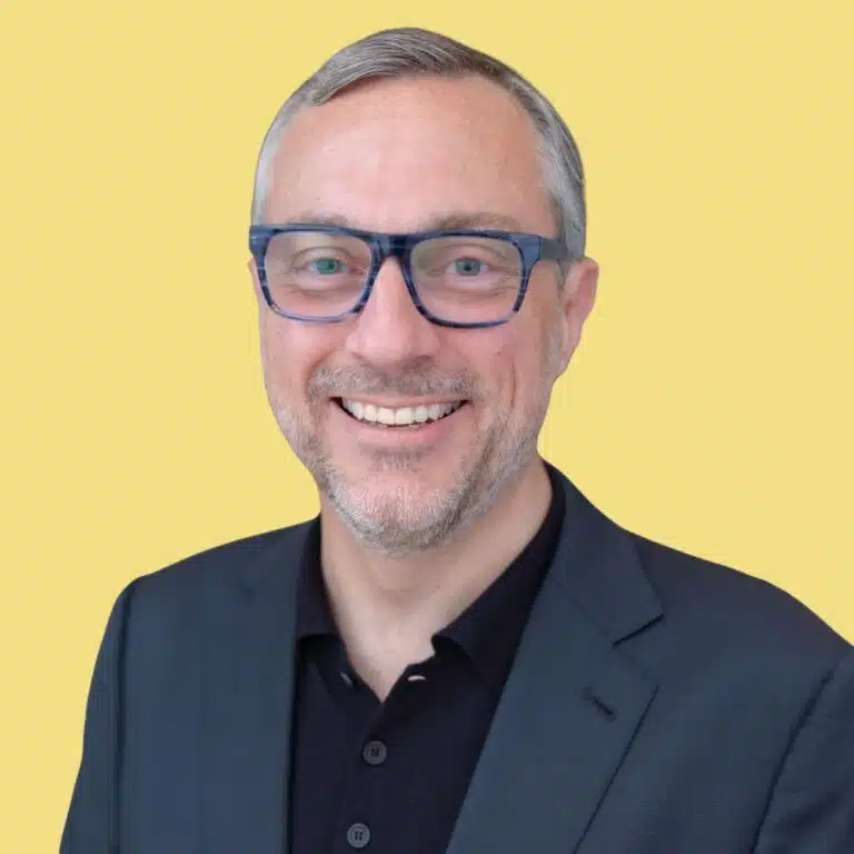 Professional headshot of man with glasses smiling against yellow background
