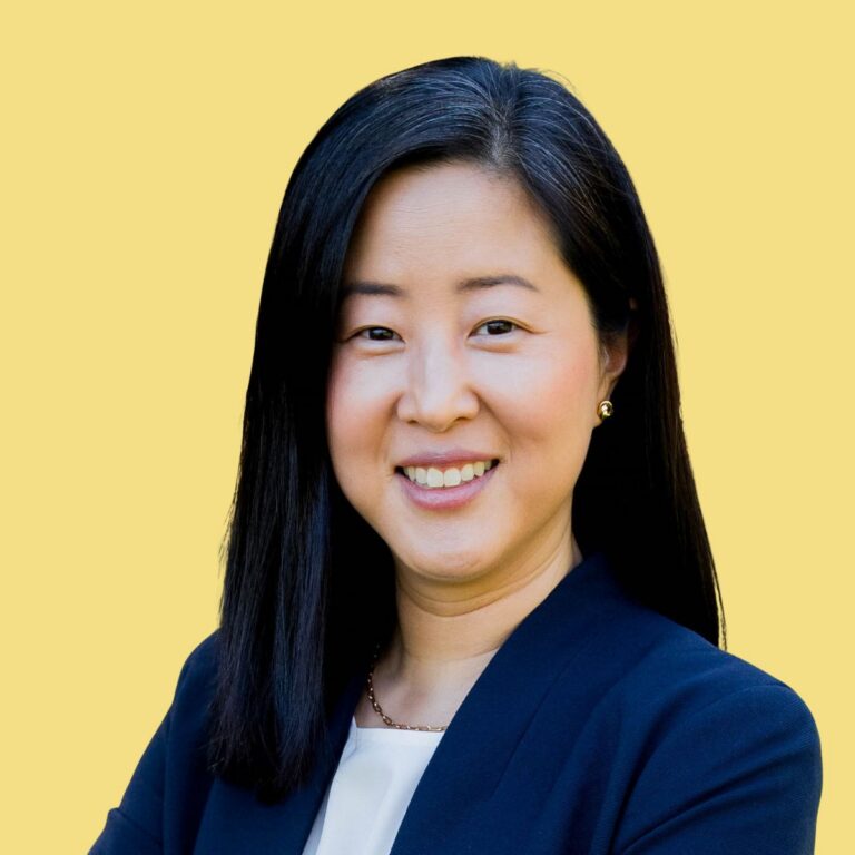 Professional headshot of Anna Moon, woman with long black hair wearing dark blazer against yellow background