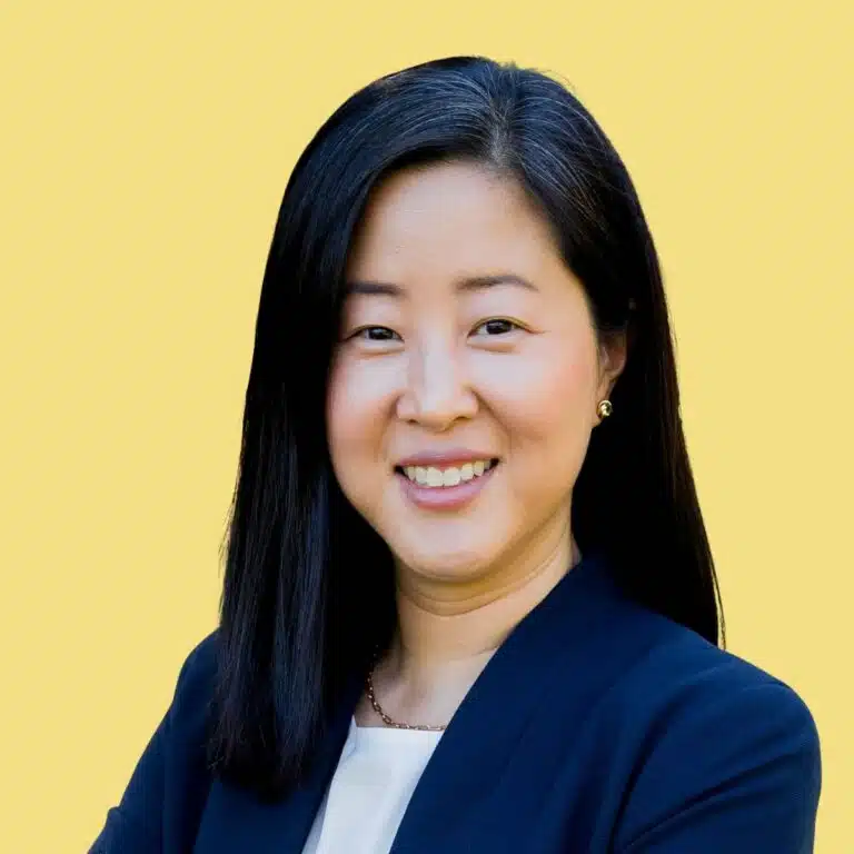 Professional headshot of Anna Moon, woman with long black hair wearing dark blazer against yellow background