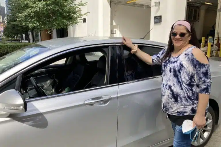 Woman in tie-dye shirt and pink visor posing beside open car door in parking garage