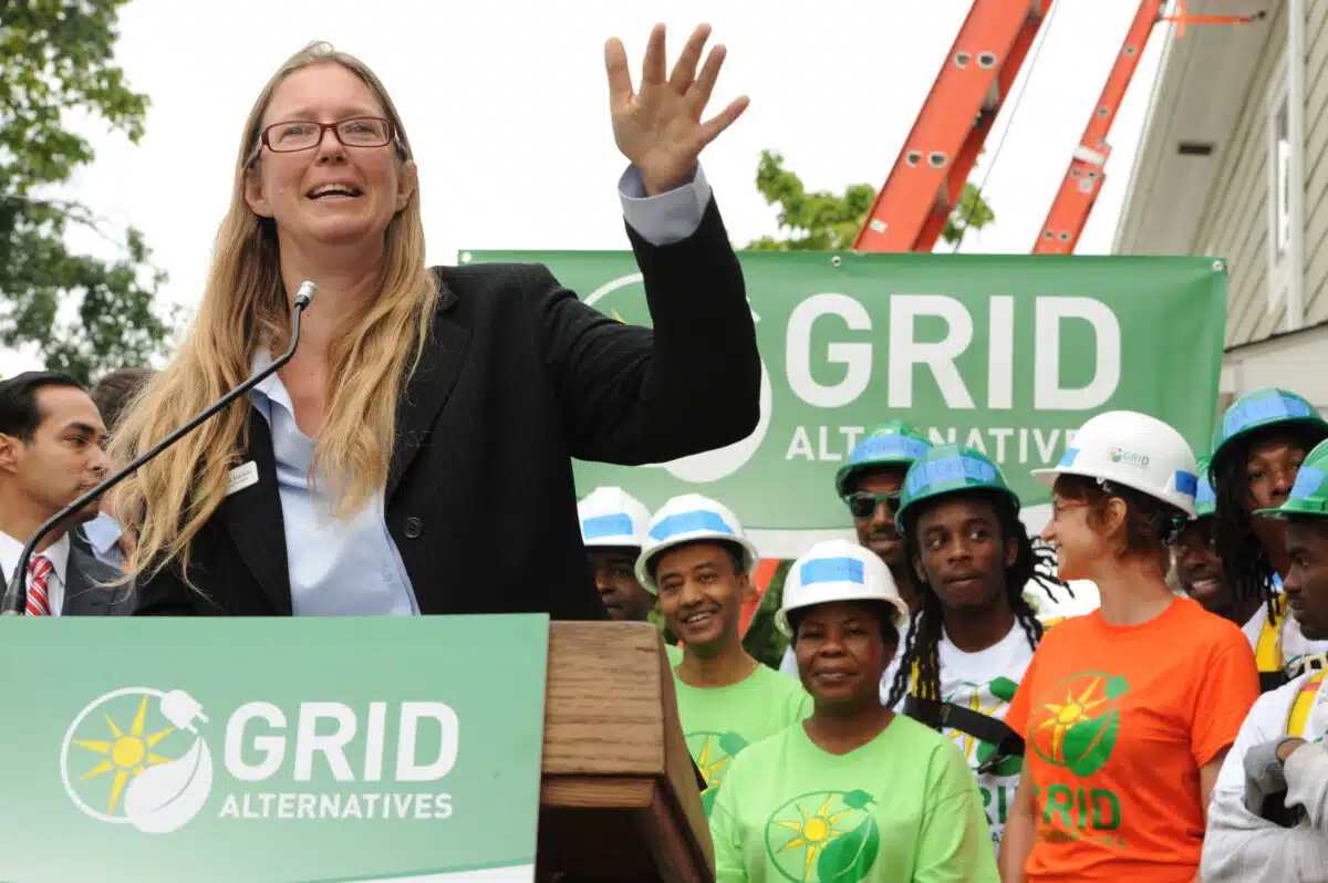 Woman speaking at podium with GRID Alternatives logo, addressing construction workers in hard hats