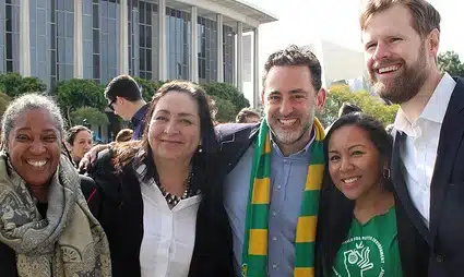 Five people smiling at outdoor event in front of building, one wearing green and gold scarf