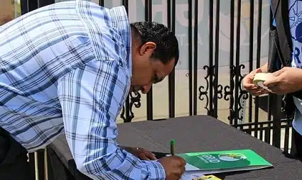Man in blue plaid shirt signing green document at table with metal fence in background
