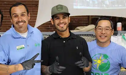 Three men posing together indoors, wearing casual clothing and smiling at the camera during what appears to be a community event