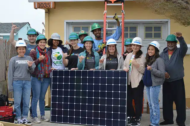 Group of people wearing hard hats posing with a solar panel in front of a yellow house numbered 11035