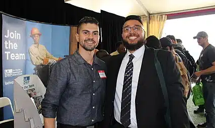 Two men smiling at a recruitment event with "Join the Team" banner visible behind them