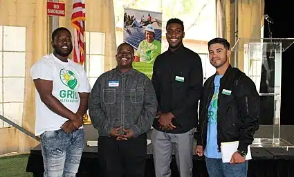 Four men smiling at camera in indoor venue, one wearing green shirt with logo, others in casual jackets and shirts