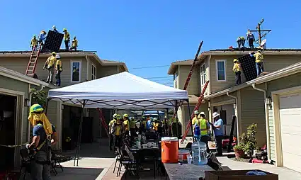 Outdoor event with white tent, firefighters on roofs, and people gathered between two houses in daylight