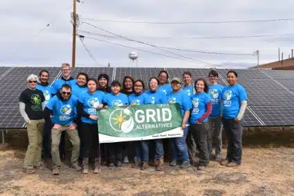 Group of volunteers in blue shirts holding GRID Alternatives banner in front of solar panels