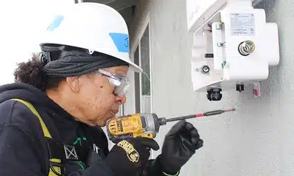 Electrician in safety helmet and glasses using multimeter to test white electrical panel on concrete wall