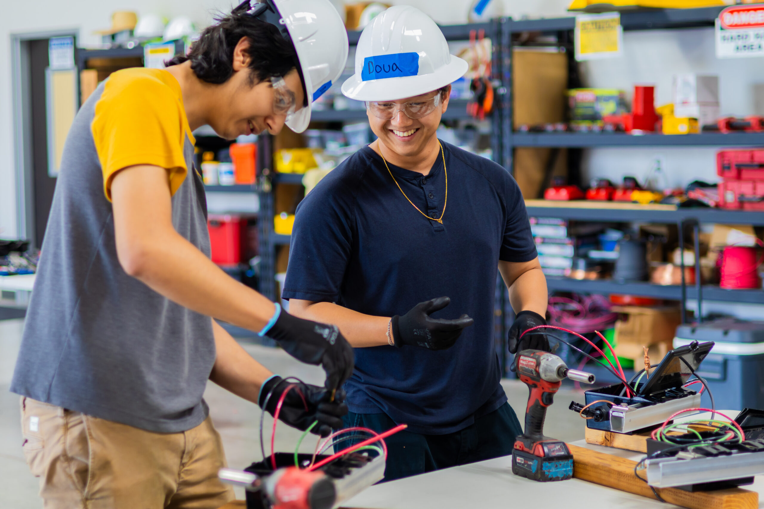 Two workers in safety helmets and gloves examine electrical components and wiring in a workshop