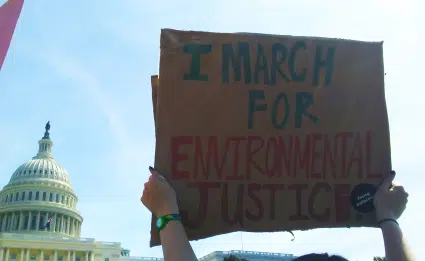 Protester holding cardboard sign reading "I march for environmental justice" with U.S. Capitol building in background